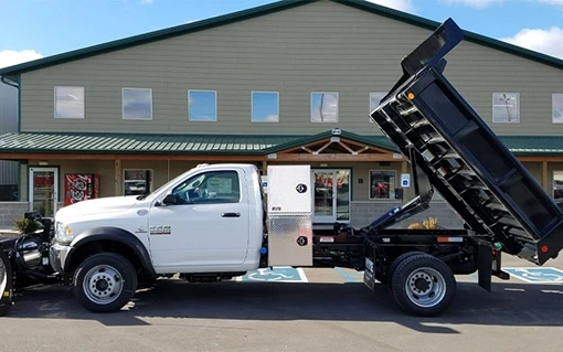 Ram Truck with Black Dump Body and Aluminum Toolboxes – MTE White Ram truck with black dump body raised and aluminum side toolboxes, viewed from the side in front of a commercial building